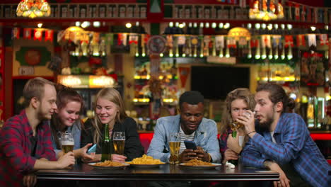 Fröhliche-Studenten-Sitzen-An-Einem-Tisch-In-Einer-Bar,-Trinken-Bier,-Essen-Chips-Und-Schauen-Sich-Fotos-Auf-Einem-Smartphone-Bildschirm-An-Und-Diskutieren-über-Fotos.